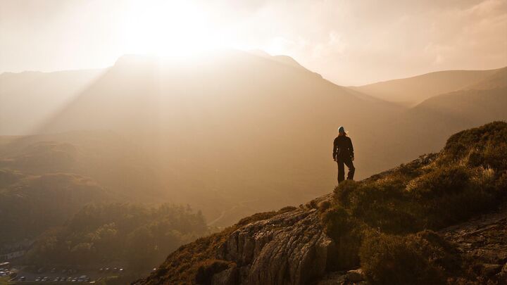 A mountainous landscape vista with the setting sun silhouetting a lone hiker on a ridge.